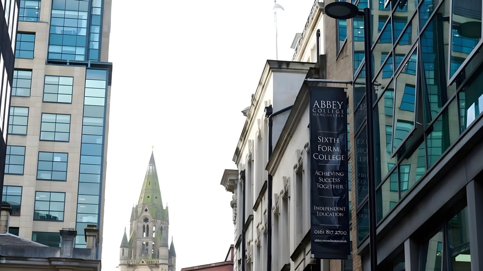 El moderno edificio de vidrio y acero del Abbey College en Manchester con una alta punta de torre de iglesia en el fondo.