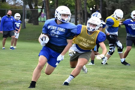 Un grupo de jugadores de fútbol de la Academy of Notre Dame entrena en un campo con árboles y edificios de fondo.
