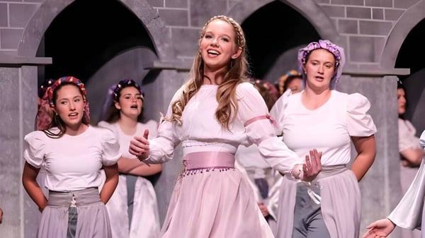 Un grupo de alumnas de la Academy of the New Church se presenta en trajes blancos y rosas en un escenario con ventanas de arco al fondo.