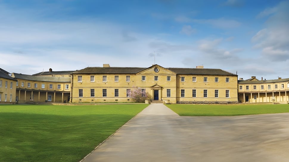 El edificio principal amarillo de la Ackworth School se encuentra en un gran campo de césped bajo un cielo azul con nubes.
