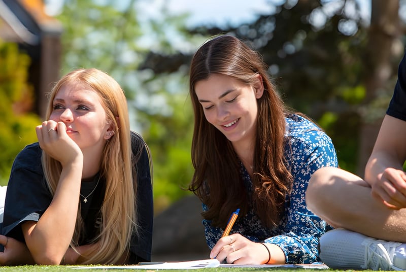 Dos alumnas están sentadas en la hierba bajo los árboles en el área exterior de la ACS International School Cobham.