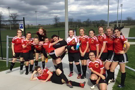 Un grupo de jugadoras de fútbol de la Acton High School posan en el campo de deportes bajo un cielo nublado.