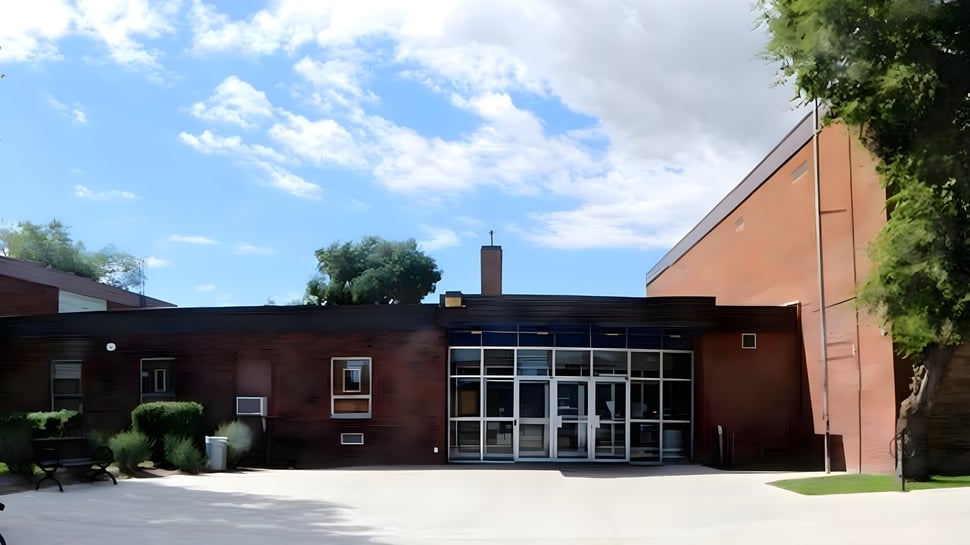 El moderno edificio de ladrillo del Aden Bowman Collegiate está rodeado de mucho verde bajo un cielo azul con nubes.