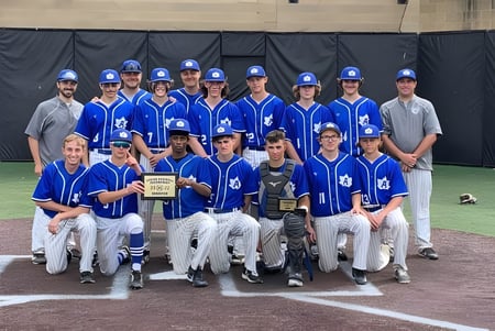 Un grupo de jugadores de béisbol en uniformes azules y blancos está en el campo de béisbol de Adrian High School.