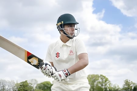 Un jugador de cricket de la Akeley Wood School está con uniforme blanco y casco en el campo frente a un cielo nublado y árboles.