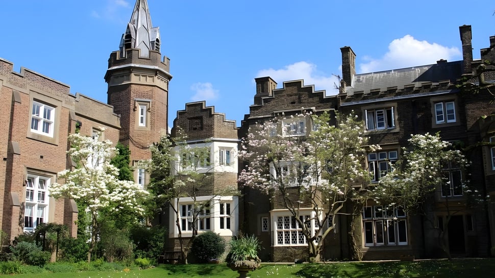 El edificio histórico de ladrillo con una alta torre se encuentra en el terreno de la Aldenham School rodeado de árboles verdes y vegetación floreciente.