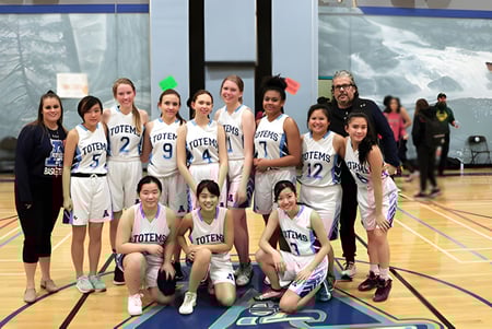 Un grupo de jóvenes estudiantes de la Aldergrove Community Secondary School posan en uniformes de baloncesto en la cancha frente a una pared de vidrio con un paisaje nevado.