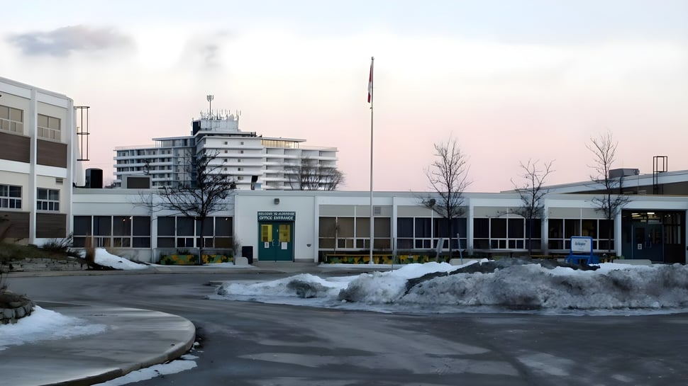 El edificio de varios pisos con una alta torre de la Aldershot High School está rodeado de un estacionamiento nevado y un cielo nublado.