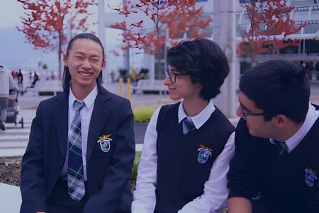 Tres alumnos en uniforme escolar están de pie frente a un edificio con árboles rojos en el campus de la Alexander Academy.