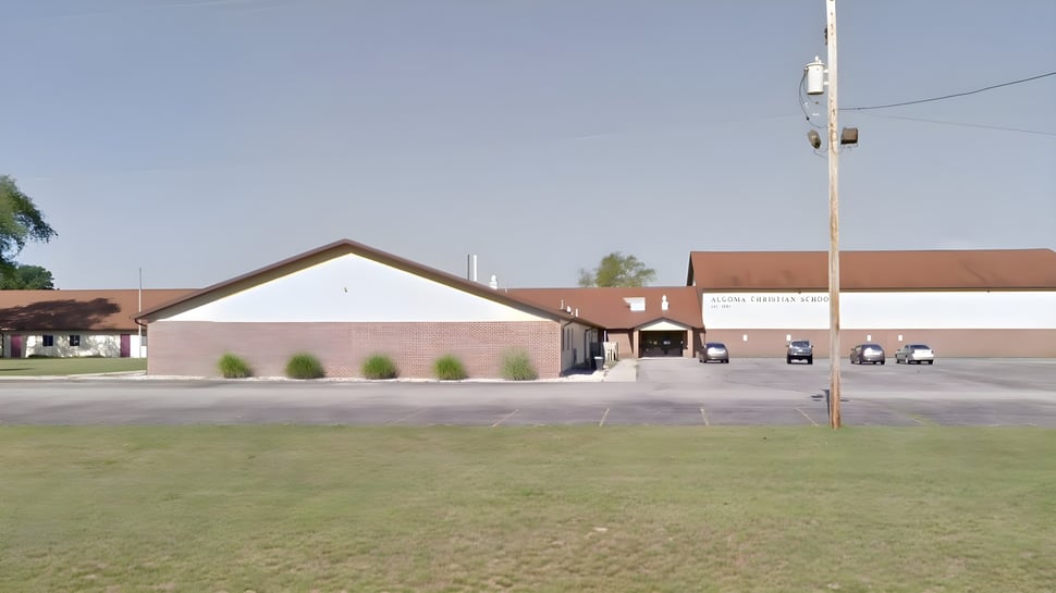 El gran edificio de ladrillo con techo a dos aguas en el campus de la Algoma Christian High School está rodeado de un prado y algunos árboles.