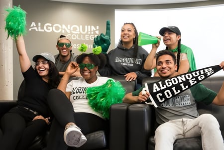Un grupo de estudiantes frente al cartel de Algonquin College sostiene pompones verdes y posa juntos.
