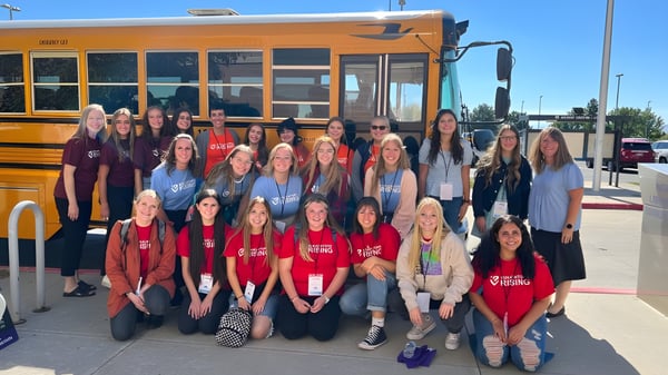 Un grupo de estudiantes del Alpine School District está frente a un autobús escolar amarillo con cielo azul de fondo.