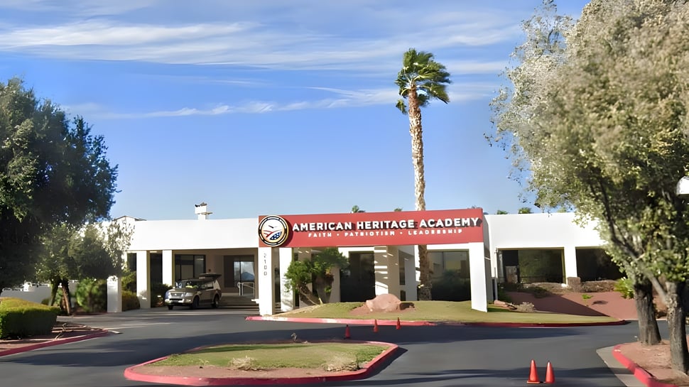 El edificio de la American Heritage Academy con un cartel rojo y palmeras en primer plano bajo un cielo azul claro.
