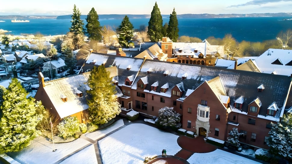 El histórico edificio de ladrillo rojo de Annie Wright Schools está rodeado de árboles cubiertos de nieve y se encuentra cerca de un cuerpo de agua con montañas al fondo.