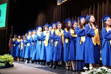 Un grupo de graduados de la Apple Valley Christian School está de pie en togas azules en un escenario frente a un logo verde.