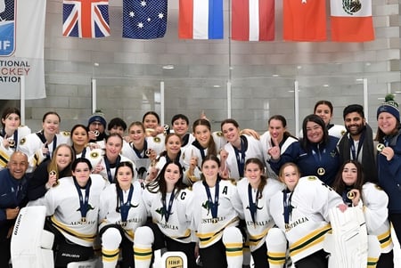 Un grupo de jugadoras de hockey sobre hielo de la Applecross Senior High School posan frente a una arena con banderas internacionales.