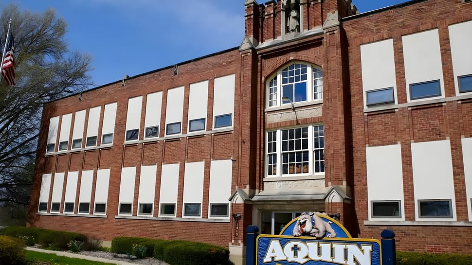 El edificio de ladrillo de varios pisos de Aquin High School está rodeado de árboles y muestra una bandera americana.