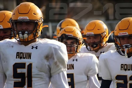 Un grupo de jugadores de fútbol de Aquin High School lleva camisetas blancas y está de pie juntos en el campo.