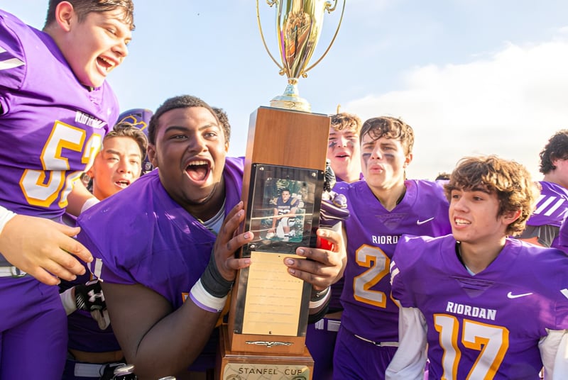 Estudiantes de la Archbishop Riordan High School celebran con un trofeo frente a un estadio bajo un cielo azul.