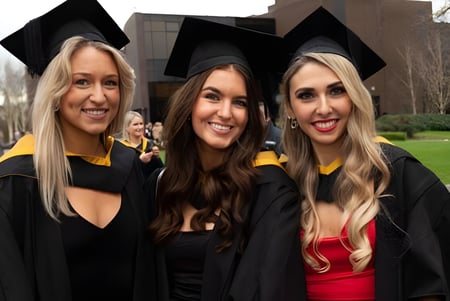 Tres graduadas de la Ardscoil Mhuire están con sombreros y togas frente a un edificio universitario.