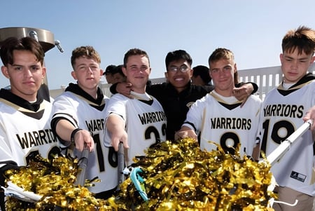 Un grupo de jóvenes atletas de la Army and Navy Academy está al aire libre en uniformes deportivos blancos y negros con la inscripción Warriors y sostiene pompones dorados.