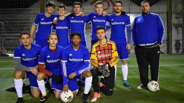 El equipo de fútbol del Ashbourne College posando en camisetas azules para una foto de equipo en el campo deportivo por la noche.