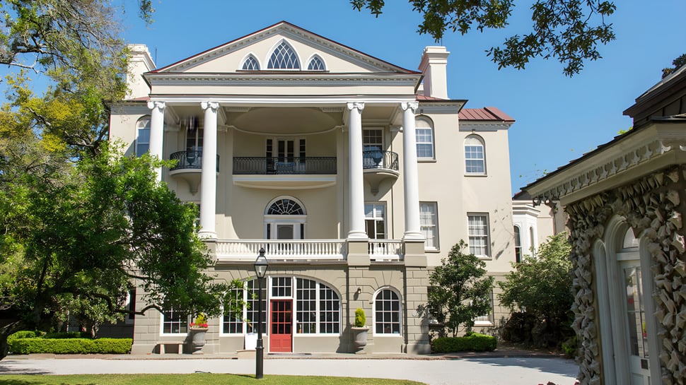 El edificio histórico con columnas y balcones en el campus de la Ashley Hall School está rodeado de árboles y vegetación verde.