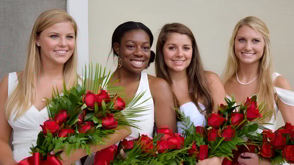 Cuatro jóvenes mujeres en vestidos blancos con ramos de rosas rojas frente a una pared blanca en el terreno de la Ashley Hall School.