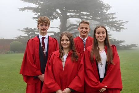 Un grupo de cuatro graduados en togas rojas está de pie en un campo en el campus del Ashville College.