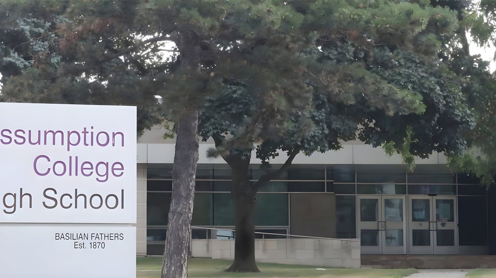 La vista exterior de Assumption College Catholic High School con un cartel y una fila de árboles en el fondo.
