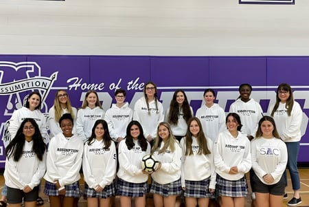 Un grupo de estudiantes en uniformes blancos está frente a una pancarta morada en el campus de Assumption College Catholic High School.