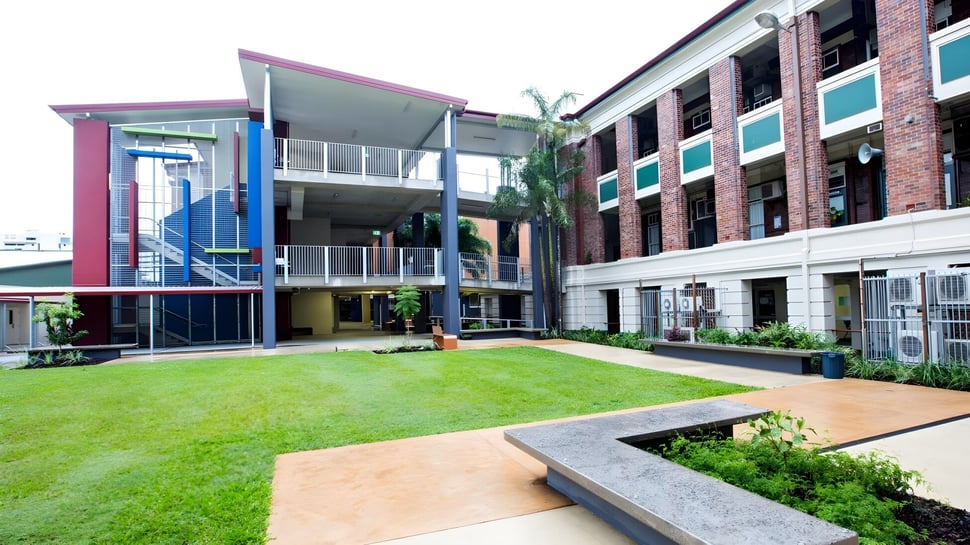 El moderno y colorido edificio de la Atherton State High School con un patio cubierto de hierba y una palmera en primer plano.