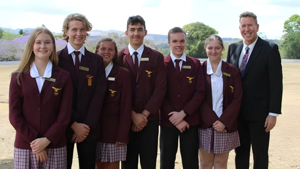 Un grupo de estudiantes de la Atherton State High School está de pie en un campo cubierto de hierba frente a edificios y árboles.