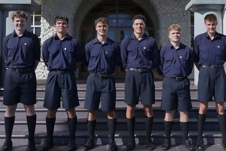 Un grupo de seis estudiantes en uniformes azules está frente a un edificio de piedra con ventanas de arco en el campus de la Auckland Grammar School.