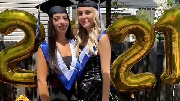 Dos estudiantes de la A.Y. Jackson Secondary School posan con ropa de graduación frente a globos dorados con el número '22'.