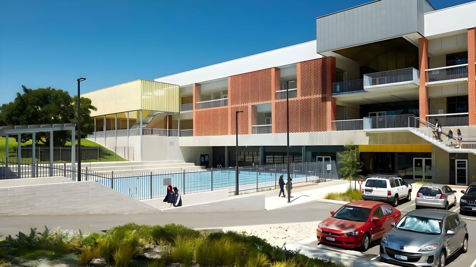 El edificio de varios pisos de la Balcatta Senior High School con una piscina en primer plano y coches estacionados está rodeado de mucho verde.