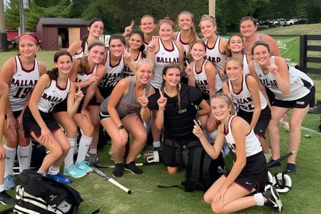 Un grupo de jóvenes atletas de la Ballard High School está de pie juntos en un campo de césped frente a un edificio y árboles.