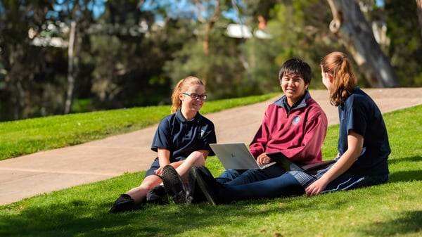 Tres estudiantes de la Banksia Park International High School están sentados en un prado con árboles de fondo.