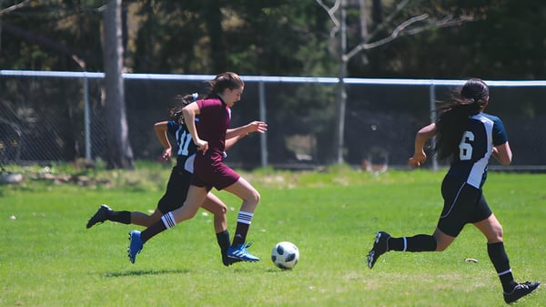 Dos futbolistas corren en el campo deportivo de la Banting Memorial High School con árboles y una cerca de fondo.
