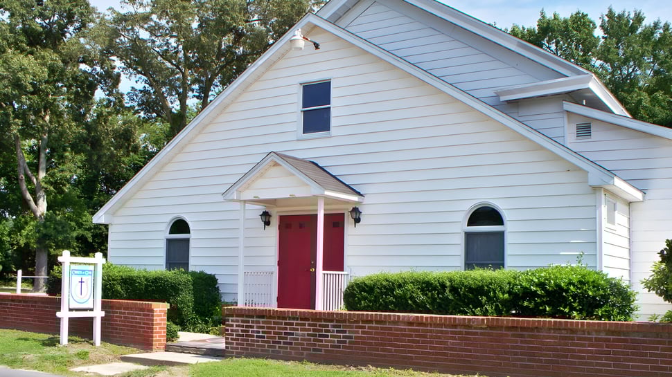 El edificio de la iglesia blanca con puerta roja y ventanas de arco en el terreno de la Beacon of Hope Christian School está rodeado de árboles verdes.