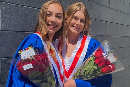Dos graduadas en coloridos trajes de graduación con ramos de flores frente a una pared de concreto gris en la Bear Creek Secondary School.