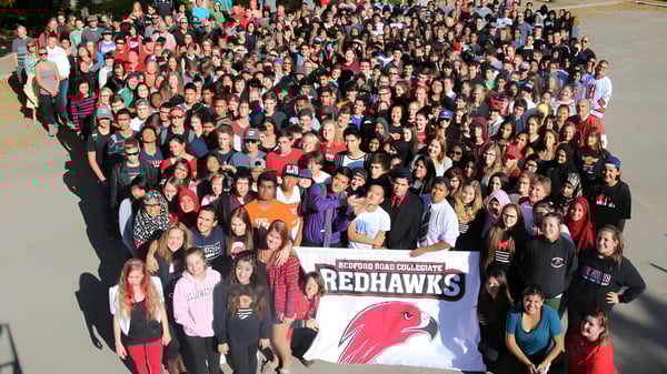Muchos estudiantes sostienen en el campus de Bedford Road Collegiate un banner con la mascota Redhawks.