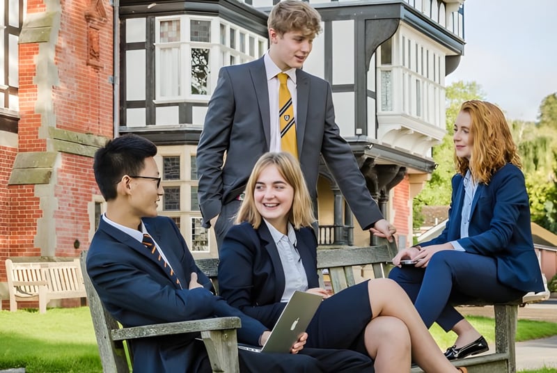 Un grupo de estudiantes está sentado en un banco al aire libre en el campus de Bedstone College frente a un edificio de ladrillo.