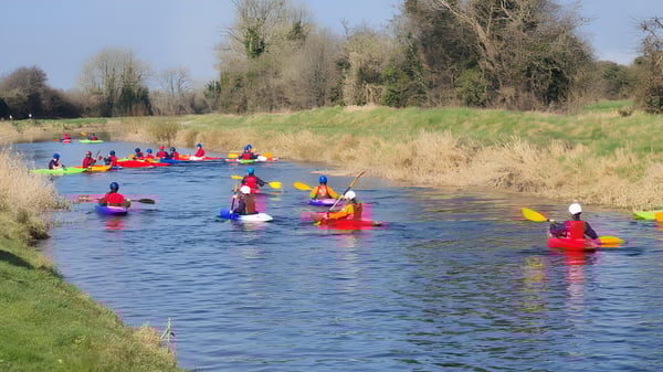 Estudiantes del Beech Hill College reman en coloridos kayaks en un río con vegetación ribereña verde.