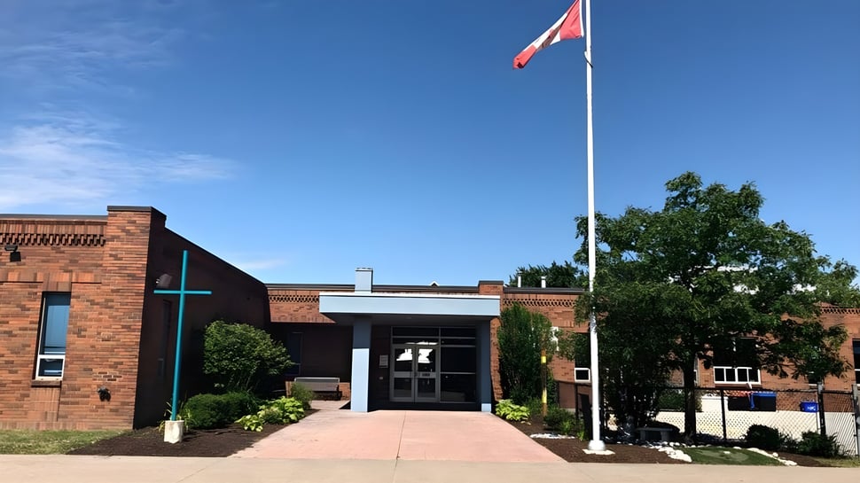 El edificio de ladrillo de la Belle River District High School con una gran bandera canadiense sobre la entrada y árboles en primer plano.