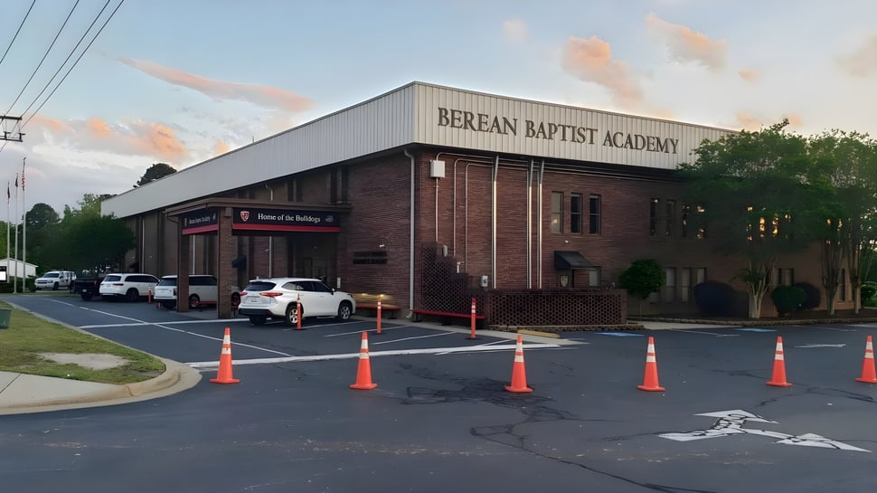 El edificio de ladrillo de la Berean Baptist Academy está rodeado de un estacionamiento con vehículos y barreras.
