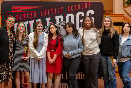 Un grupo de estudiantes se encuentra frente a un banner de la Berean Baptist Academy con diferentes estilos de ropa.