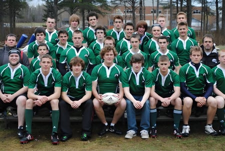 Un grupo de estudiantes de la Bernice McNaughton High School está sentado en uniformes deportivos en un campo frente a un bosque.