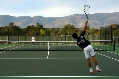Una persona con ropa deportiva juega al tenis en el campo de tenis de la Besant Hill School con montañas y vegetación de fondo.