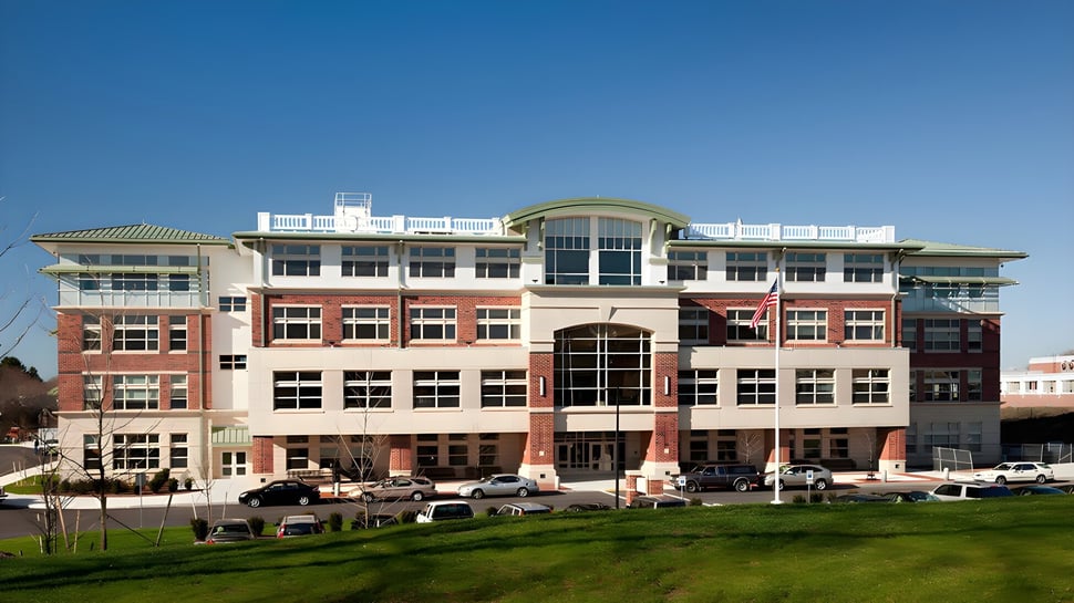 El edificio de varios pisos de la Beverly High School está rodeado de un área verde y coches estacionados bajo un cielo azul claro.